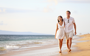 A couple walks hand in hand on the beach, with gentle waves lapping at the shore under a bright sky.