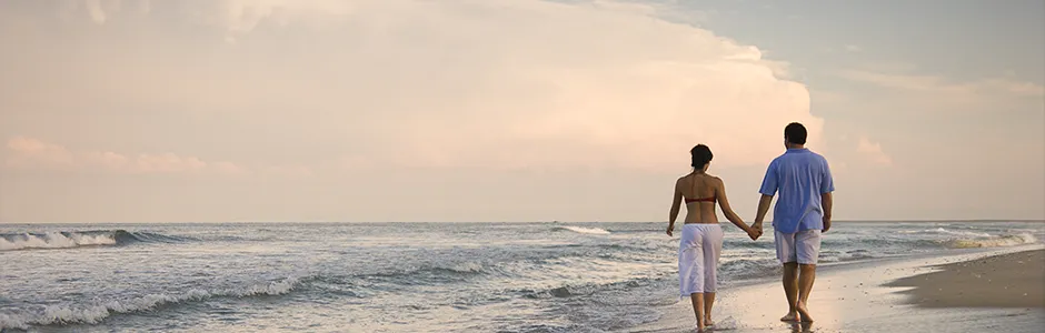 A couple strolls hand in hand along a sandy beach, with gentle waves lapping at the shore in the background.