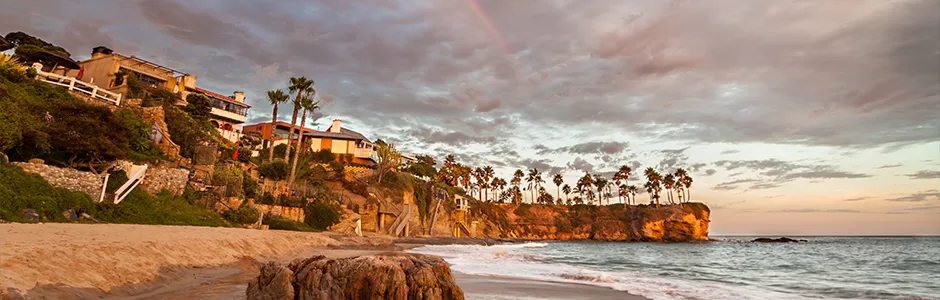 A scenic beach with soft waves and a vibrant rainbow arching across a clear blue sky.